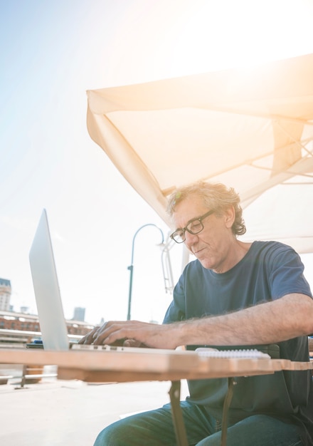 Älterer Mann, der am caf at im Freien schreibt auf Laptop sitzt