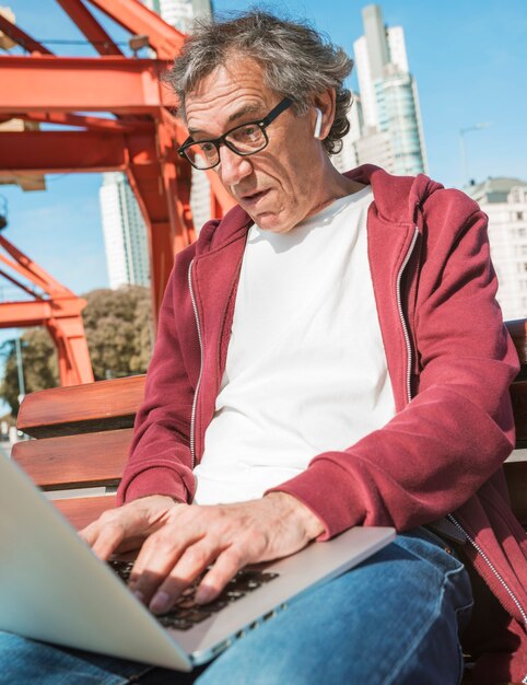 Älterer Mann, der auf Bank unter Verwendung des Laptops an draußen sitzt