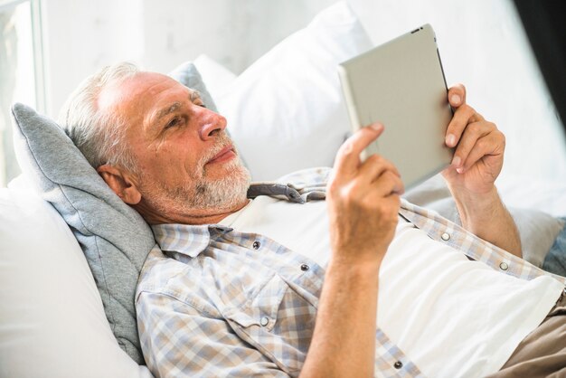 Älterer Mann, der auf dem Bett betrachtet digitale Tablette liegt