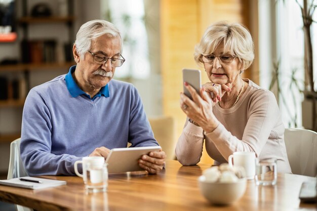 Älterer Mann, der auf dem Touchpad im Internet surft, während seine Frau zu Hause SMS auf dem Handy sendet