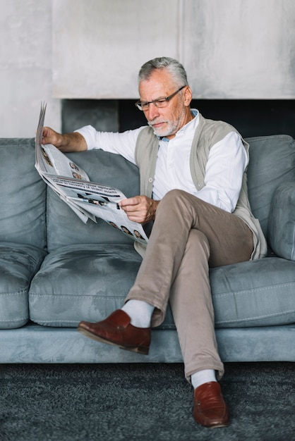 Älterer Mann, der auf gemütlichem Sofa mit gekreuzter Beinlesezeitung sitzt