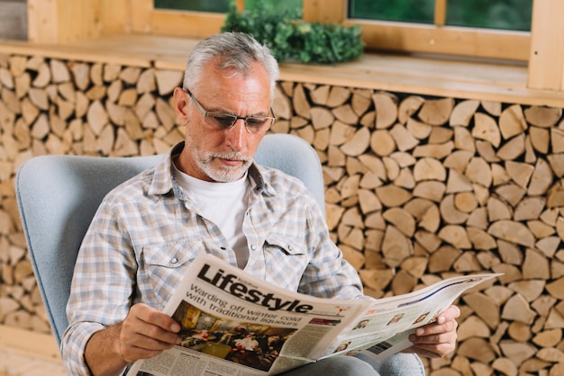 Älterer Mann, der auf Stuhllesezeitung nahe dem Fenster sitzt