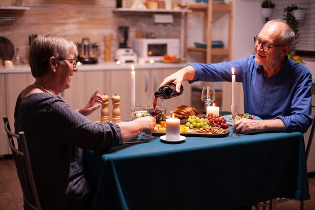 Älterer Mann, der Frau Wein einschenkt, während er Beziehungsjubiläum in der Küche feiert. Romantisches Paar sitzt am Tisch im Esszimmer, redet, genießt das Essen.