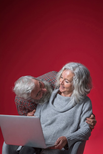 Älterer Mann, der ihre Frau sitzt auf dem Stuhl hält einen offenen Laptop gegen roten Hintergrund betrachtet