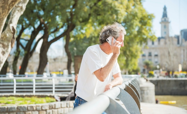 Älterer Mann, der nahe dem Geländer spricht am Handy im Park steht