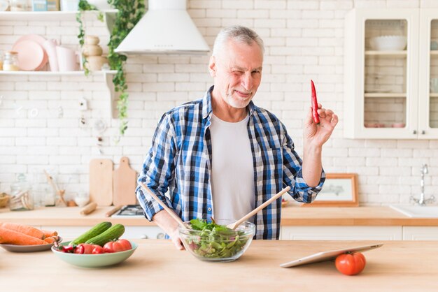 Älterer Mann, der sein Auge blinzelt, das in der Hand den Pfeffer des roten Paprikas zubereitet den Salat zubereitet