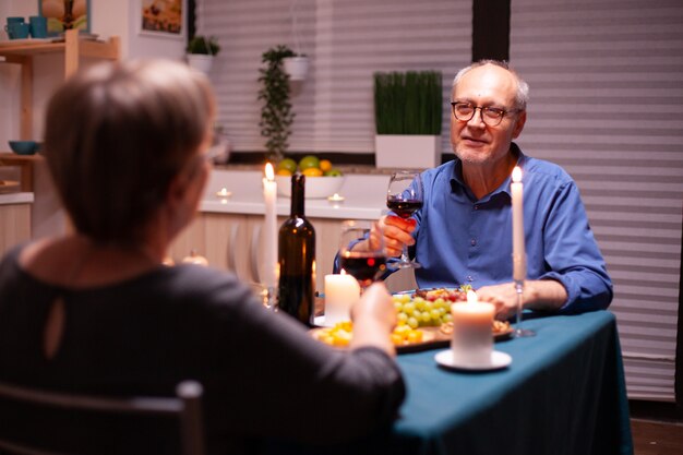 Älterer Mann, der seiner Frau eine Geschichte erzählt, während er in der Küche mit Wein und Essen feiert. Älteres Paar sitzt am Tisch im Esszimmer, redet, genießt das Essen, feiert ihr Jubiläum in