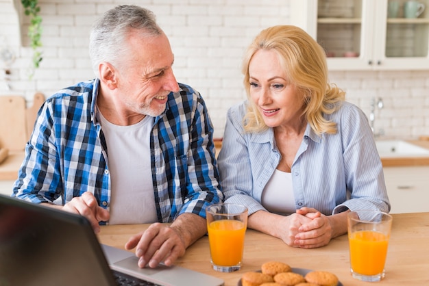 Älterer Mann, der seiner Frau etwas auf Laptop mit Glassaft und Muffins auf Tabelle zeigt
