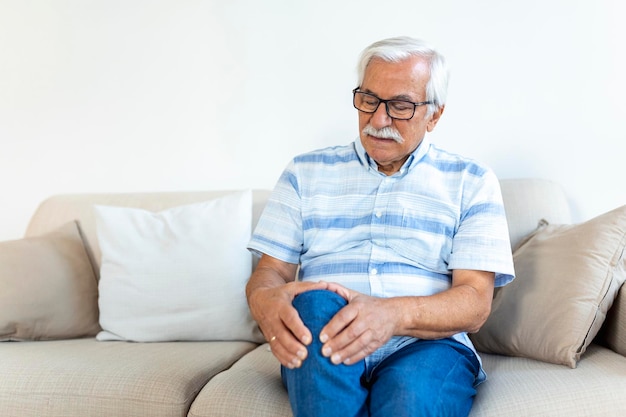 Älterer Mann, der zu Hause auf einem Sofa sitzt und sein schmerzhaftes Knie berührt Menschengesundheitsversorgung und Problemkonzept Unglücklicher älterer Mann, der zu Hause an Knieschmerzen leidet