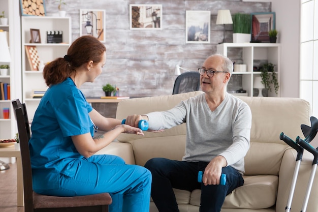 Älterer Mann im Pflegeheim mit Hanteln, der mit Hilfe der Krankenschwester Physiotherapie macht.