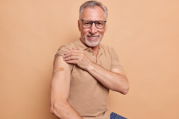 Älterer Mann in Brille zeigt verputzten Arm, nachdem er einen Coronavirus-Impfstoff erhalten hat und sich glücklich fühlt, sich sicher und geschützt isoliert über einer braunen Wand zu fühlen