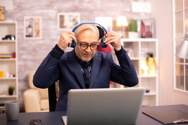 Älterer Mann in den 60ern mit Kopfhörern auf dem Kopf, der Musik hört und an einem modernen Laptop arbeitet