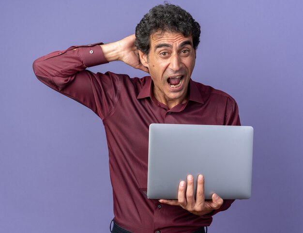 Älterer Mann in lila Hemd hält Laptop schreiend und schaut erstaunt und überrascht auf blauem Hintergrund stehend