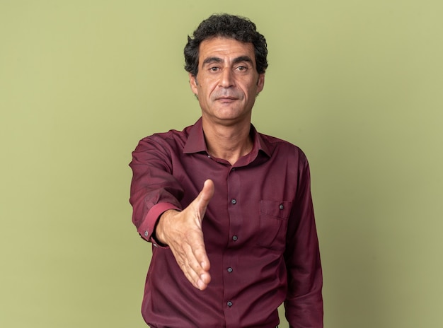 Älterer Mann in lila Hemd mit Armgrußgeste, der selbstbewusst auf grünem Hintergrund steht