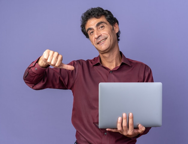 Älterer Mann in lila Hemd mit Laptop, der die Kamera anschaut und selbstbewusst auf sich selbst zeigt, der über blauem Hintergrund steht