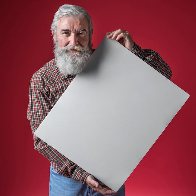 Älterer Mann mit dem grauen Bart, der in der Hand weißes leeres Plakat gegen roten Hintergrund hält