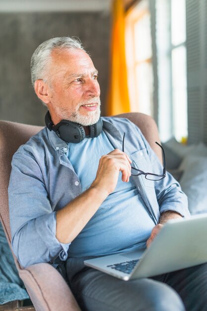Älterer Mann mit dem Laptop, der auf dem Lehnsessel weg schaut sitzt