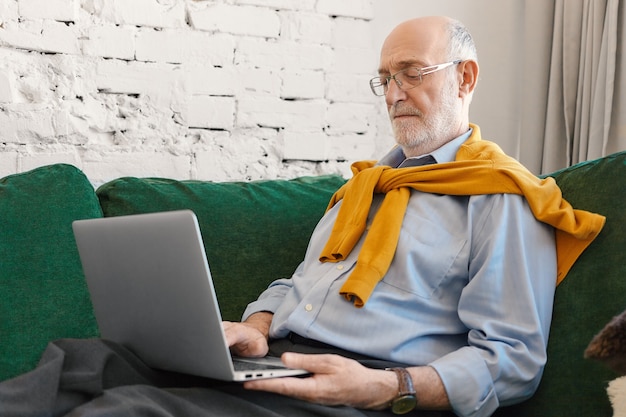 Älterer Mann mit Glatze und Bart unter Verwendung der drahtlosen Hochgeschwindigkeits-Internetverbindung zu Hause auf Laptop. Ernsthafter konzentrierter reifer Geschäftsmann, der Geschäftsnachrichten auf tragbarem Computer auf Couch liest