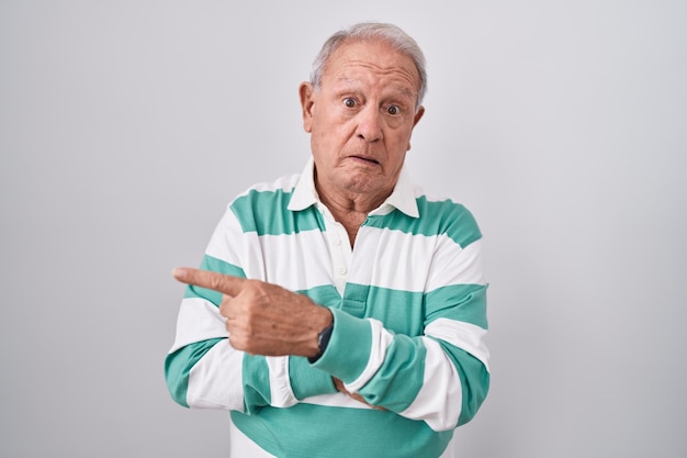 Älterer Mann mit grauen Haaren steht vor weißem Hintergrund, zeigt beiseite, besorgt und nervös, mit beunruhigtem und überraschtem Zeigefinger.