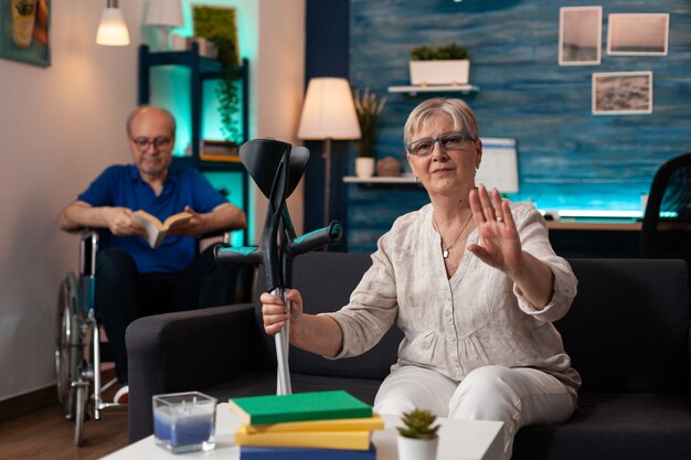 Älterer Mann und Frau mit Krücken und Rollstuhl sitzen zu Hause auf der Couch. Kaukasischer alter Mann liest ein Buch und eine Frau winkt mit der Hand in die Kamera auf Videoanruf-Technologiegerät-Gadget