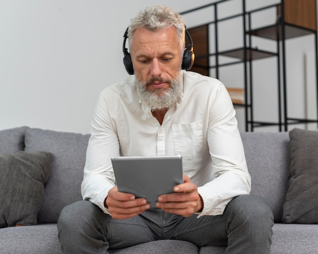 Älterer Mann zu Hause auf der Couch mit Tablet-Gerät und Kopfhörern