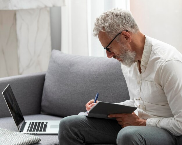 Älterer Mann zu Hause, der am Laptop lernt und Notizen macht