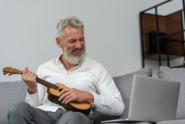 Älterer Mann zu Hause, der Ukulele-Unterricht auf dem Laptop studiert