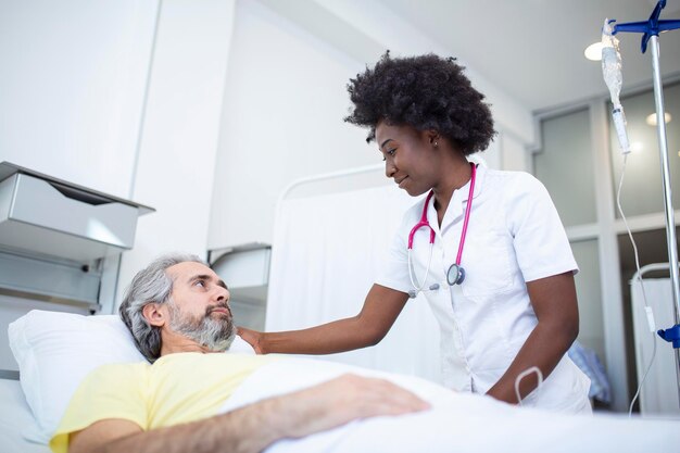 Älterer Patient im Bett, der mit afroamerikanischer Ärztin im Krankenzimmer spricht Gesundheits- und Versicherungskonzept Arzt, der ältere Patienten im Krankenhausbett tröstet oder Diagnosegesundheit berät