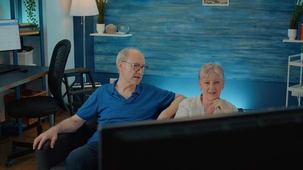 Älteres Ehepaar sieht sich Filme im Fernsehen an, um den Ruhestand im Wohnzimmer zu genießen und Spaß mit Comedy im Fernsehen zu haben. Senioren sitzen auf dem Sofa und schauen sich den Comedy-Kanal auf dem Bildschirm an, um Spaß zu haben.