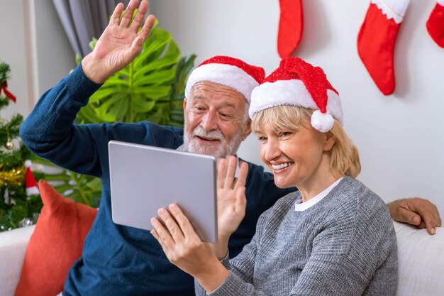 Älteres Paar, das ein Tablet zum Videoanruf nutzt, um seine Familie zum Weihnachtsfest zu begrüßen, winkt mit der Hand auf dem Sofa mit Dekoration und Baum
