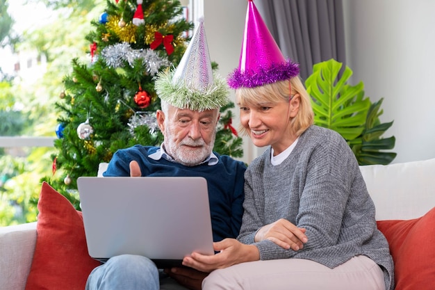 Älteres Paar, das Laptop zum Videoanruf verwendet, um seine Familie zum Weihnachtsfest zu begrüßen, das auf Sofa mit Dekoration und Baum sitzt