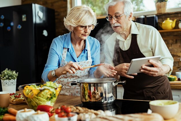 Älteres Paar kocht, während es dem Rezept auf dem Touchpad in der Küche folgt