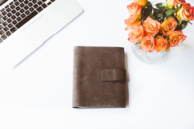 Luftaufnahme eines weißen schreibtisches mit einem mit leder bezogenen notizbuch, einem laptop und einer blumenvase