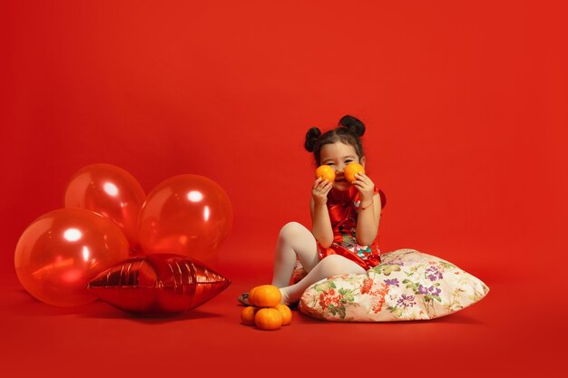 Luftballons und Mandarinen für Stimmung. Positiv posieren. . Asiatisches niedliches kleines Mädchen lokalisiert auf roter Wand in traditioneller Kleidung. Feier, menschliche Gefühle, Feiertage. Copyspace.