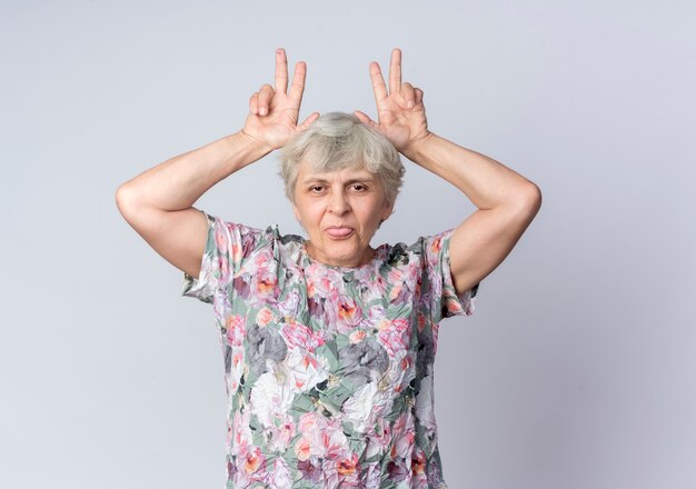 Lustige ältere Frau legt Hände auf Kopf gestikulierende Hörner und streckt Zunge heraus, die auf weißer Wand isoliert wird