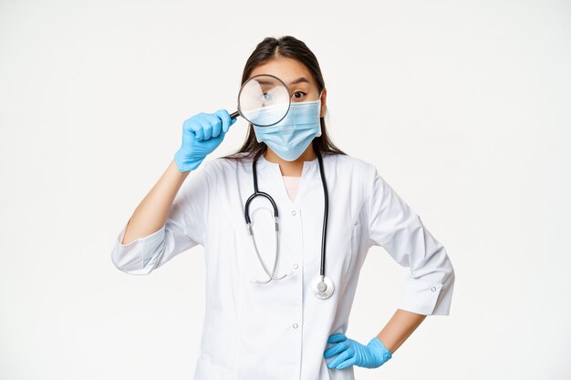 Lustige asiatische Ärztin schaut durch eine Lupe auf den Patienten, trägt eine medizinische Gesichtsmaske und Gummihandschuhe, weißer Hintergrund