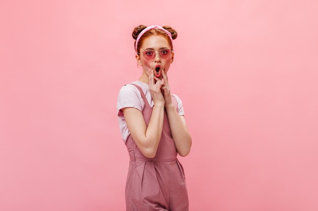 Lustige Frau mit Brötchen macht Gesicht. Frau im rosa Outfit und in den Gläsern, die auf lokalisiertem Hintergrund aufwerfen.