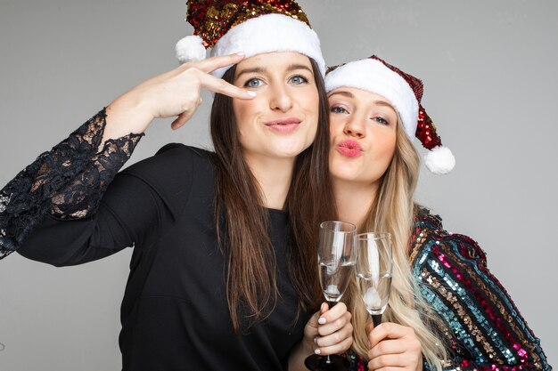 Lustige Frauen in Kleidern und Weihnachtsmützen trinken Champagner und posieren für die Kamera