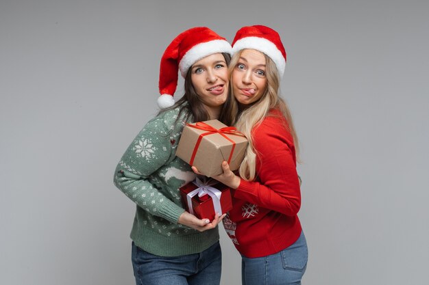 Lustige Frauen in rot-weißen Weihnachtsmützen halten sich gegenseitig Geschenke und posieren für die Kamera