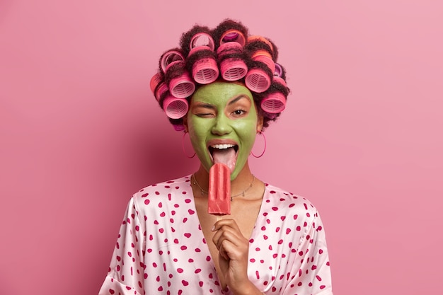 Lustige freudige Hausfrau leckt leckeres Eis, zwinkert Auge, genießt das Essen von kaltem erfrischendem Sommerdessert, trägt grüne Maske im Gesicht, Lockenwickler, in lässigem Outfit gekleidet, hat Spaß zu Hause