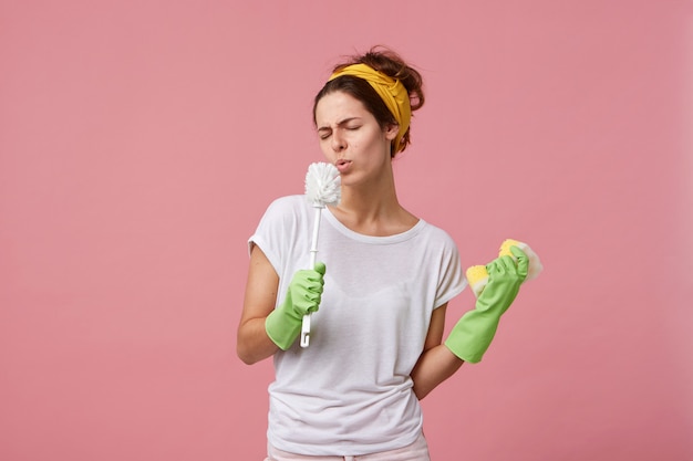 Lustige junge europäische Hausfrau, die Stirnband, lässiges T-Shirt und grüne Gummihandschuhe trägt, die Reinigungsprozess zu Hause genießen, Schwamm und Toilettenbürste wie ein Mikrofon an ihrem Mund halten und unterschreiben