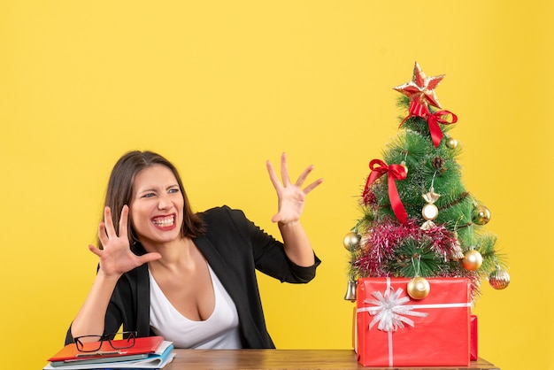 Lustige junge Frau, die etwas mit überraschtem Gesichtsausdruck betrachtet, der an einem Tisch nahe geschmücktem Weihnachtsbaum im Büro auf Gelb sitzt