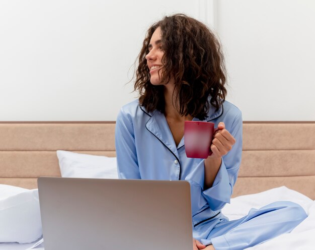 Lustige junge schöne Frau in den blauen Pyjamas, die auf Bett mit Tasse Kaffee arbeiten, der auf Laptop glücklich und positiv schaut beiseite im Schlafzimmerinnenraum auf hellem Hintergrund arbeitet