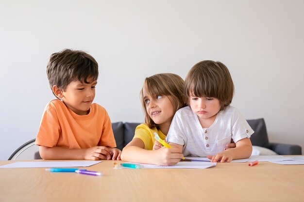 Lustige Kinder, die mit Markierungen im Wohnzimmer malen. Schönes blondes Mädchen, das Bruder ansieht. Kinder sitzen am Tisch, zeichnen mit Stiften und spielen zu Hause. Kindheit, Kreativität und Wochenendkonzept