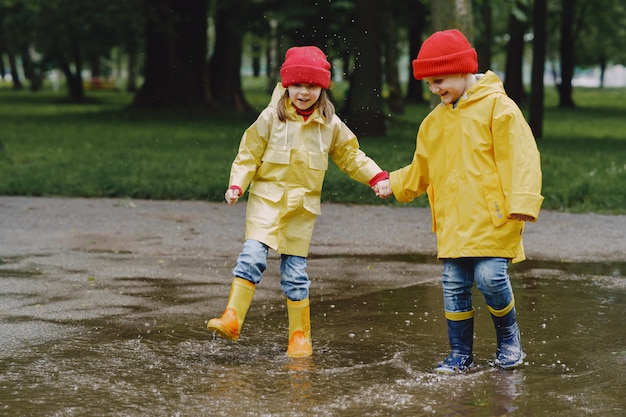 Lustige Kinder in Regenstiefeln, die mit Papierschiff durch eine Pfütze spielen