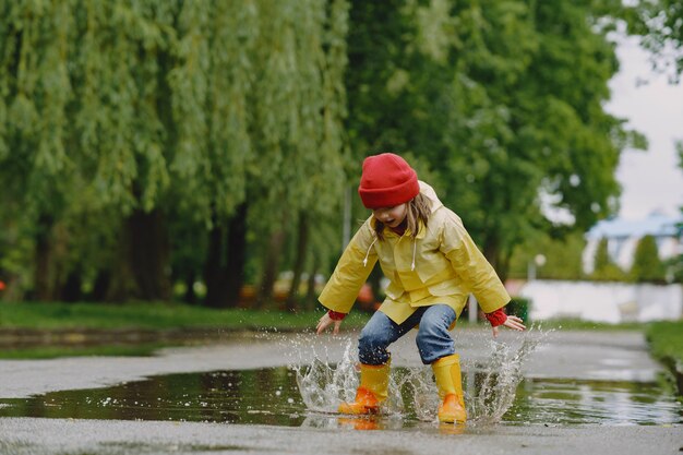 Lustige Kinder in Regenstiefeln, die mit Papierschiff durch eine Pfütze spielen