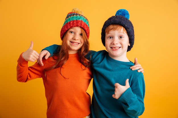 Lustige kleine Kinder, welche die warmen Hüte sich zeigen Daumen tragen.