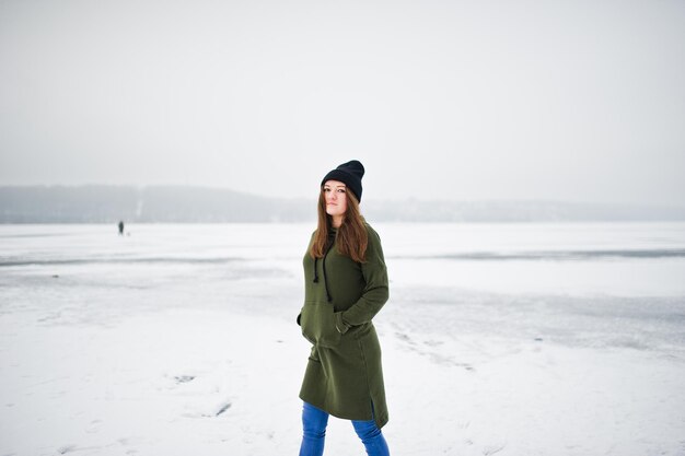 Lustige Mädchen tragen lange grüne Sweatshirt-Jeans und schwarze Kopfbedeckungen am zugefrorenen See im Wintertag