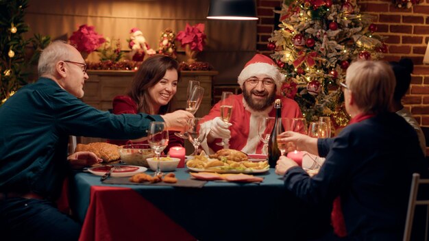 Lustige Person, die als Weihnachtsmann kostümiert ist und sich mit der Familie über Geschenke unterhält, während sie das Weihnachtsessen zu Hause genießt. Festlicher Mann mit traditioneller Tracht, der Winterurlaub mit Verwandten feiert.