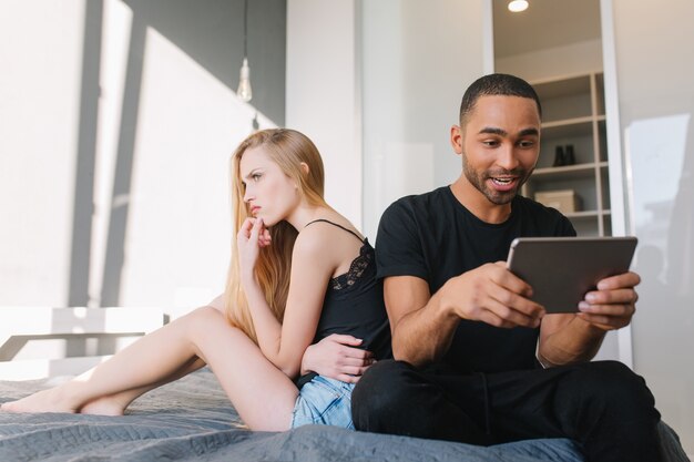 Lustige schöne Momente zu Hause der attraktiven verärgerten lustigen jungen Frau, die Fenster auf Bett nahe aufgeregtem glücklichem Kerl spielt, der mit Tablette spielt. Beziehung, beleidigtes Mädchen, Spaß haben